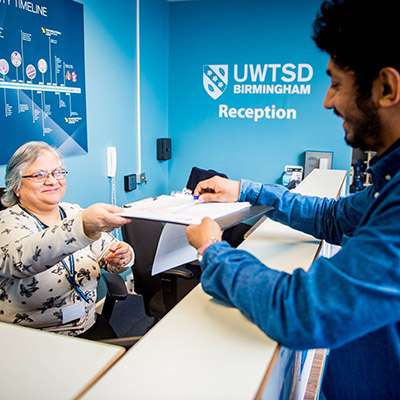 A smiling UWTSD receptionist receives a clipboard from a student.