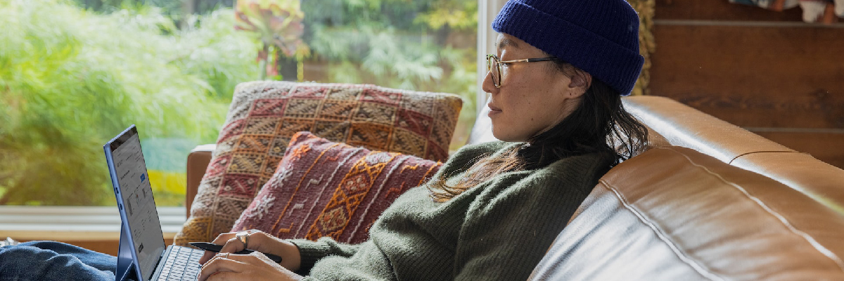 一名戴着蓝色帽子、穿着深绿色羊毛套头衫的女子靠在沙发上，在笔记本电脑上打字，看上去很放松。
