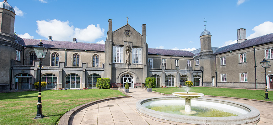 A fountain in the grassy quadrangle of Coleg Dewi Sant. The path around the fountain leads up to the main entrance of the old stone building.