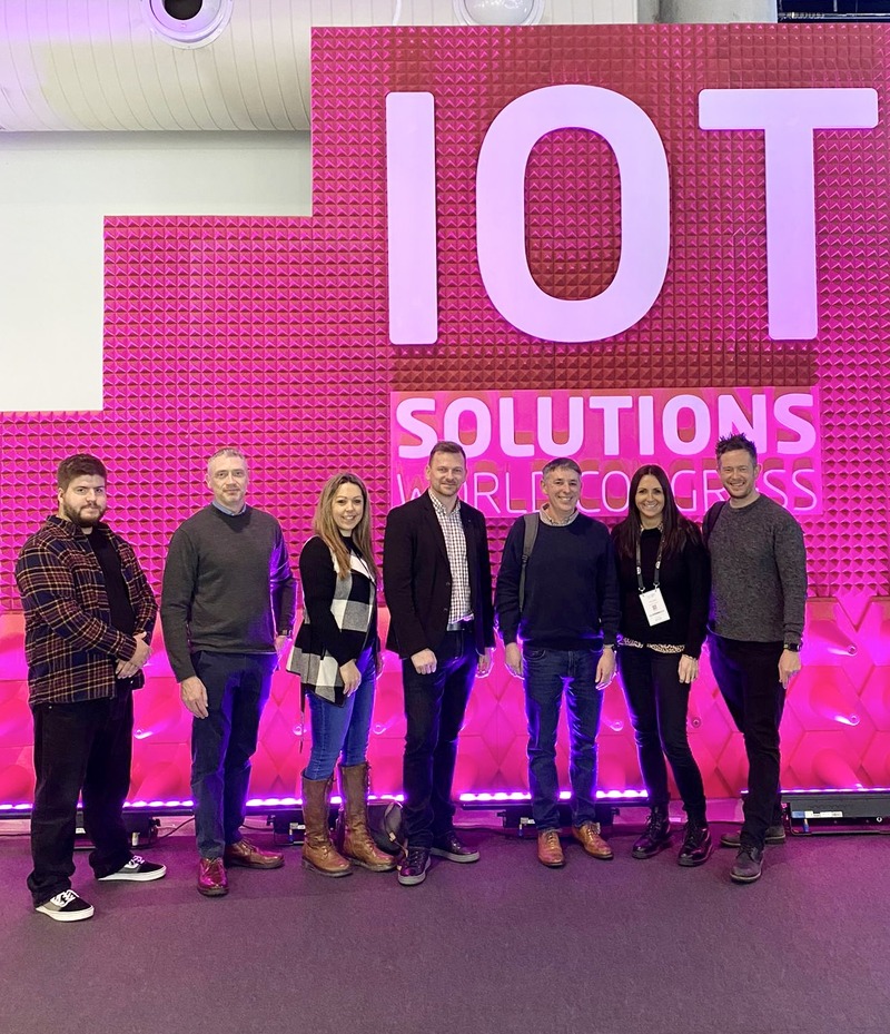 Seven members of the delegation stand smiling in front of the hot pink glowing IOT solutions sign.