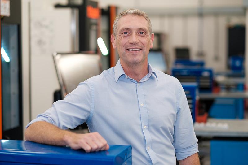 In shirtsleeves, Richard Morgan stands smiling in a room full of equipment, one arm resting on a blue metal box.