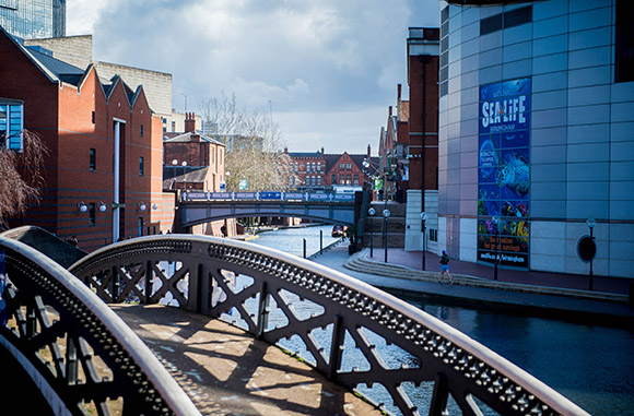 A view of the restored Canal Basin in the centre of Birmingham; a jogger runs along a towpath in front of a glassy modern building.