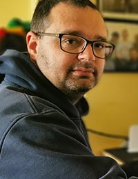 Julian Milligan, a man with glasses wearing a dark hoodie, looks over his shoulder at the camera.