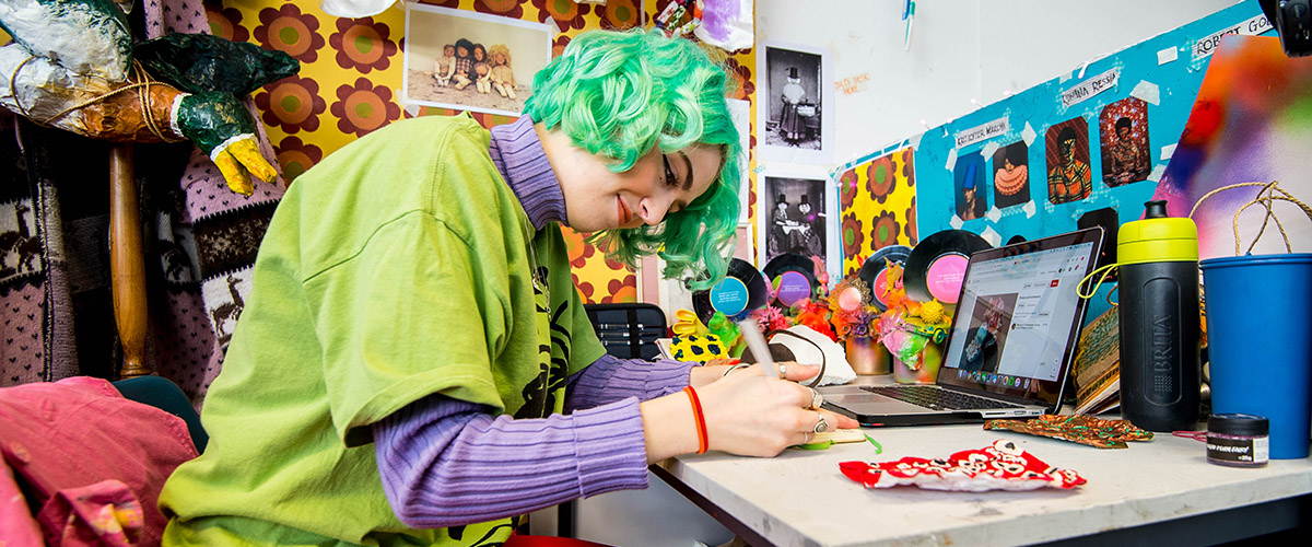 一个学生穿着色彩鲜艳的衣服，留着一头灰绿色的头发，在便利贴上写着;她桌子周围的墙壁上有鲜艳的图案;一只纸做的烤鸭挂在她身后的架子上。