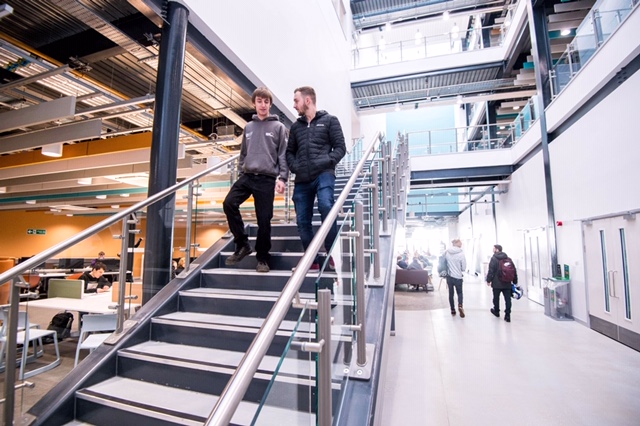 Two students walk down a flight of stairs in the metal-and-glass atrium of SA1.