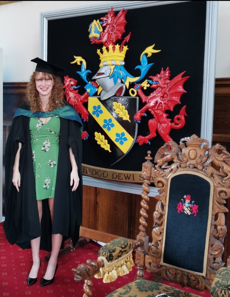 安娜·萨德勒穿着学术长袍，戴着帽子站在大学的盾徽前。