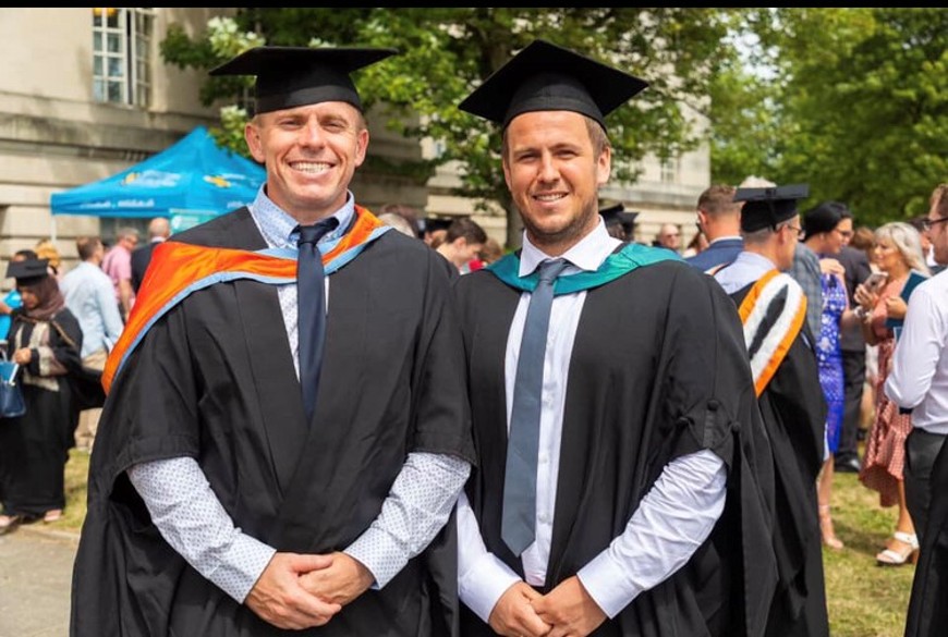 Rhys Surringer and Andrew Thomas at graduation wearing academic gowns and hats.